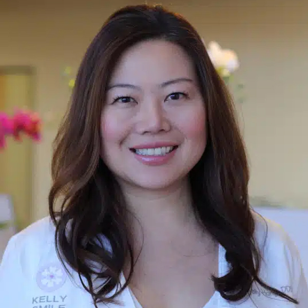 Professional woman smiling at camera in dental office setting wearing white coat with name tag.