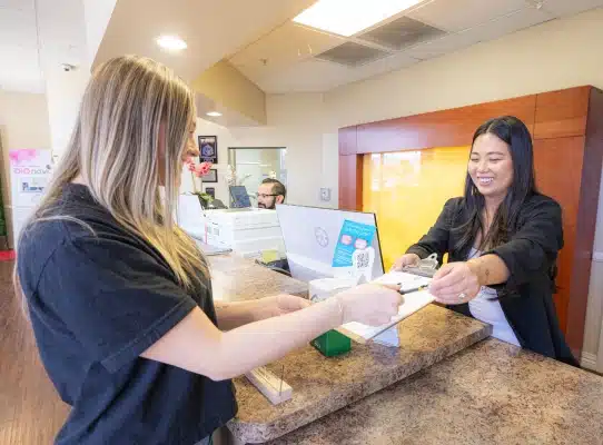 Two women at dental reception desk, one patient checking in while staff member assists in modern office.