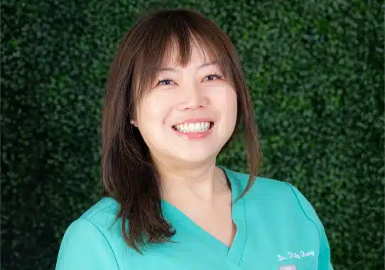 Woman in turquoise scrubs smiling in front of green foliage backdrop at dental office.