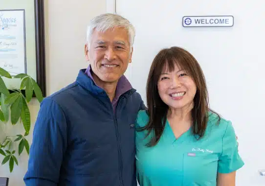 Two dental professionals smiling at camera in bright, welcoming dental office with plant and welcome sign.