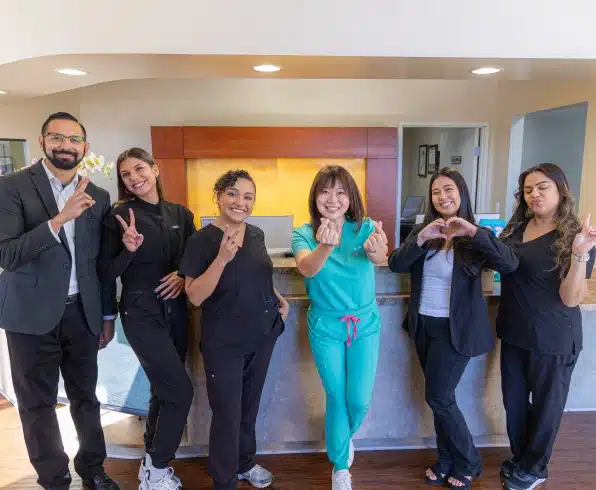 Seven dental professionals posing together in modern office hallway, giving thumbs up gestures.