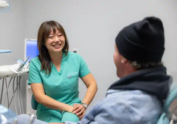 Dental hygienist in green scrubs speaking with patient in clinical treatment room.