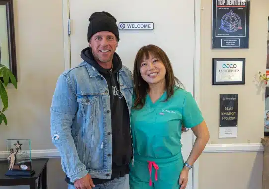 Two people posing together in a dental office reception area with certificates on wall.