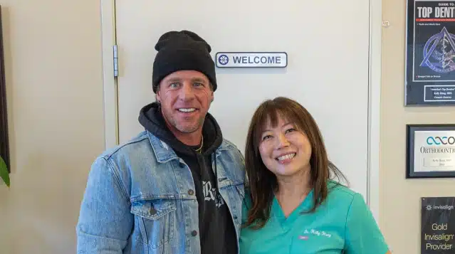 Two people smiling at dental office entrance with welcome sign visible behind them.