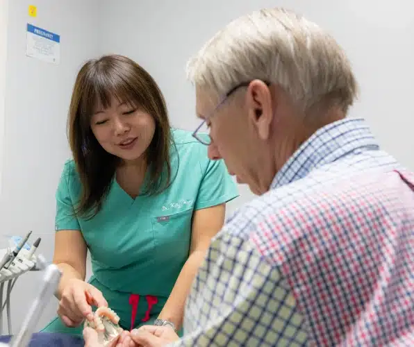 Female dentist in green scrubs demonstrating dental care to elderly male patient in clinical setting.